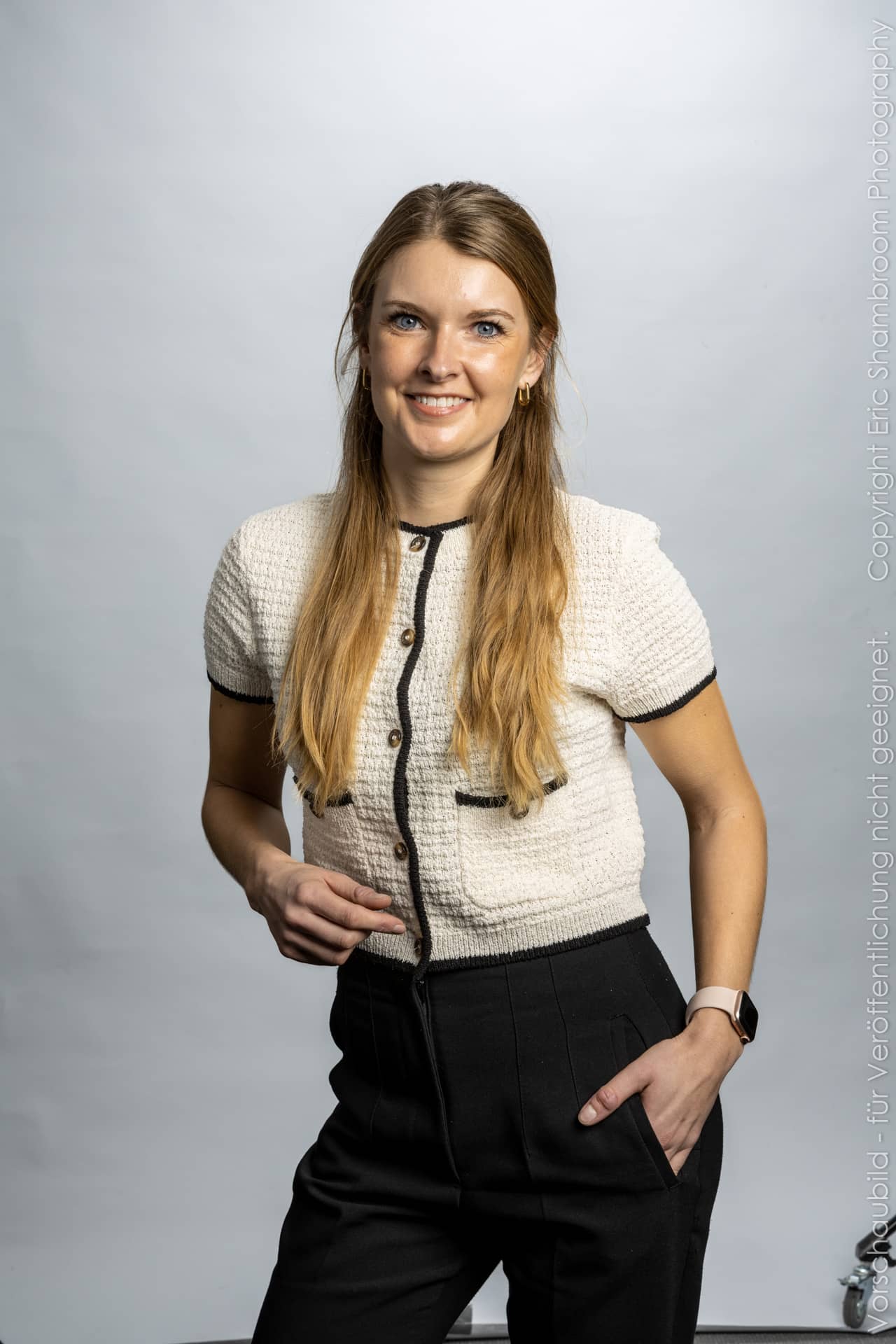 Eine lächelnde Frau mit langen, blonden Haaren trägt ein kurzes, helles Oberteil und schwarze Hose vor einem neutralen H...