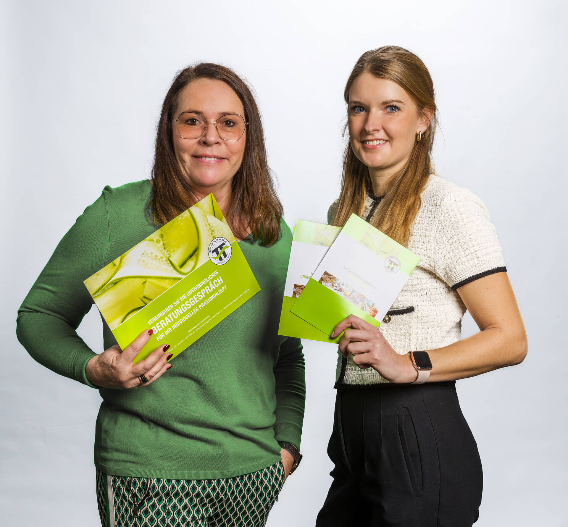 Zwei Frauen halten Informationsmaterialien in den Händen und lächeln freundlich in die Kamera.