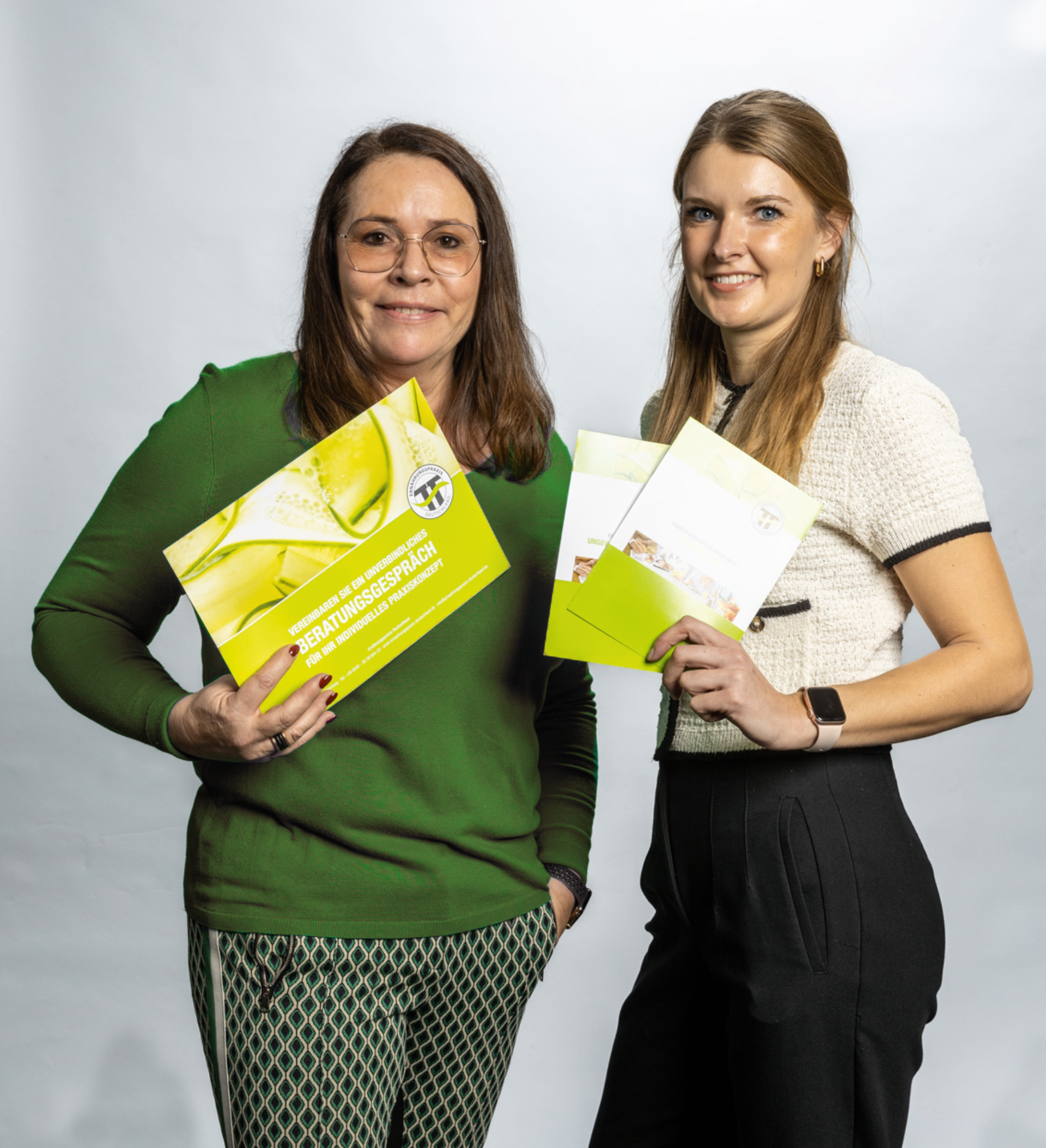 Zwei Frauen halten Informationsbroschüren in der Hand und posieren für ein Foto vor hellem Hintergrund.