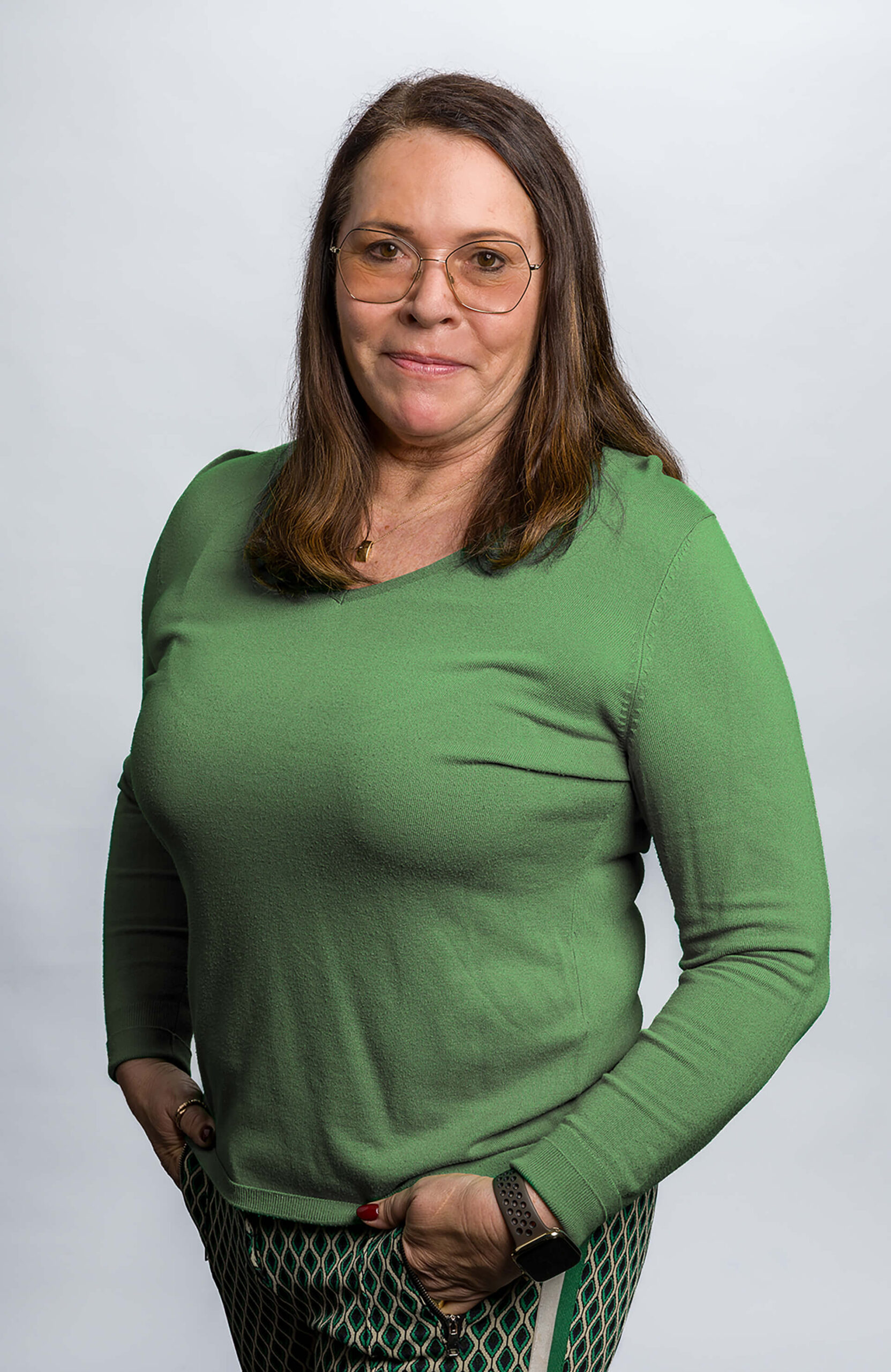 Lächelnde Frau mit langen Haaren in einem grünen Pullover und gemusterter Hose, vor neutralem Hintergrund.